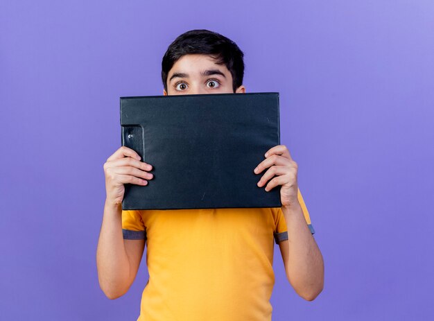 Impressed young caucasian boy holding clipboard  from behind it isolated on purple wall with copy space