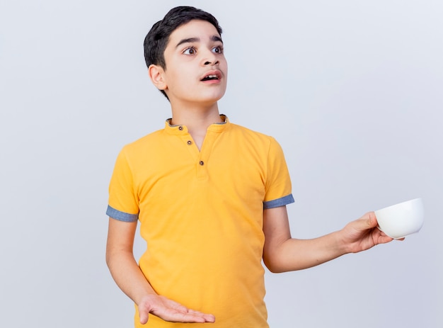 Impressed young caucasian boy holding cup showing empty hand looking at side isolated on white background with copy space