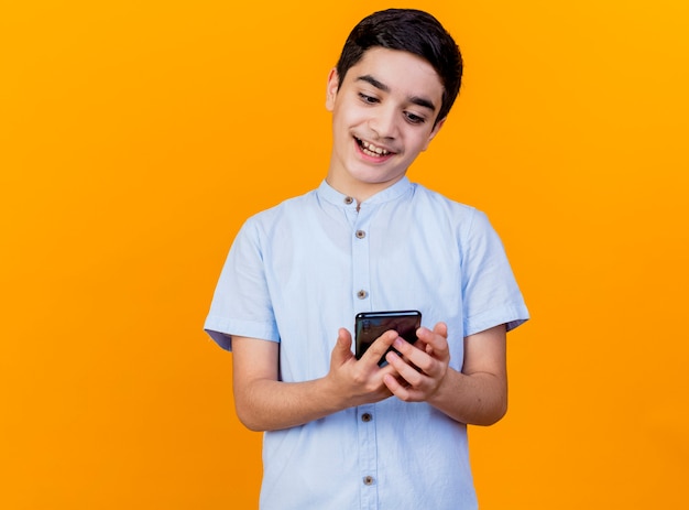 Impressionato giovane ragazzo caucasico tenendo e guardando il telefono cellulare isolato su sfondo arancione con copia spazio