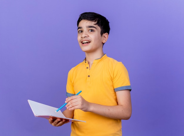 Impressed young caucasian boy holding note pad and pencil  isolated on purple wall with copy space