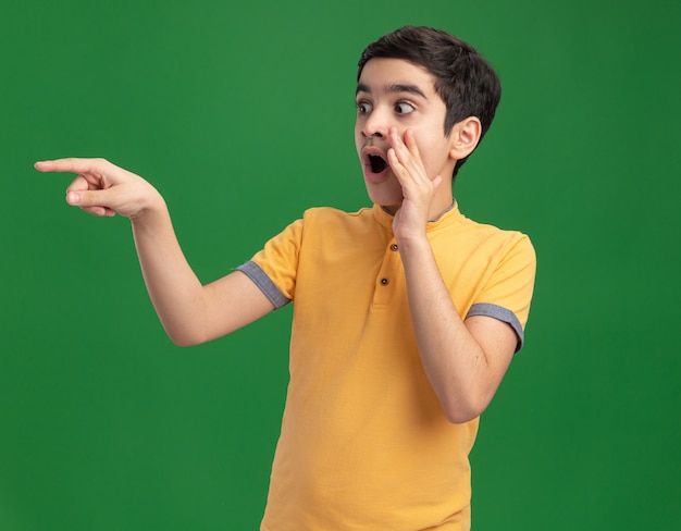 Impressed young caucasian boy keeping hand near mouth looking and pointing at side whispering 