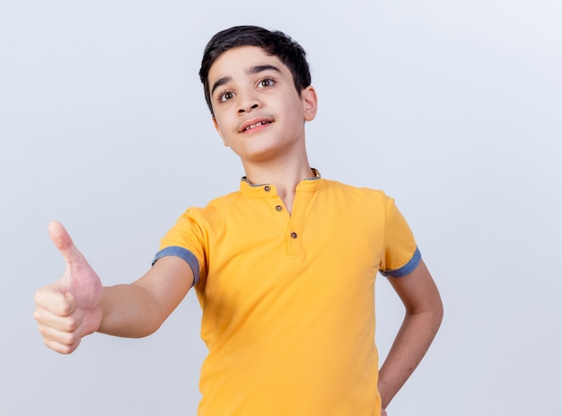 Impressed young caucasian boy keeping hand on waist looking straight showing thumb up isolated on white background with copy space