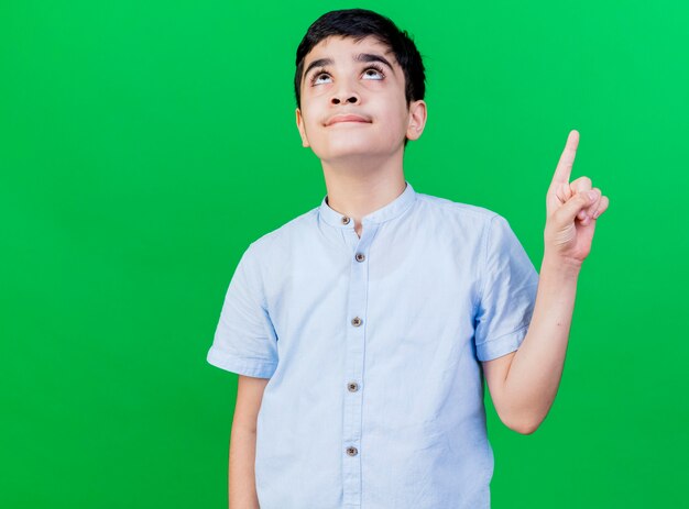 Impressed young caucasian boy looking and pointing up isolated on green wall with copy space