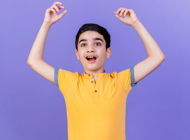 Impressed young caucasian boy looking straight raising up hands isolated on purple wall