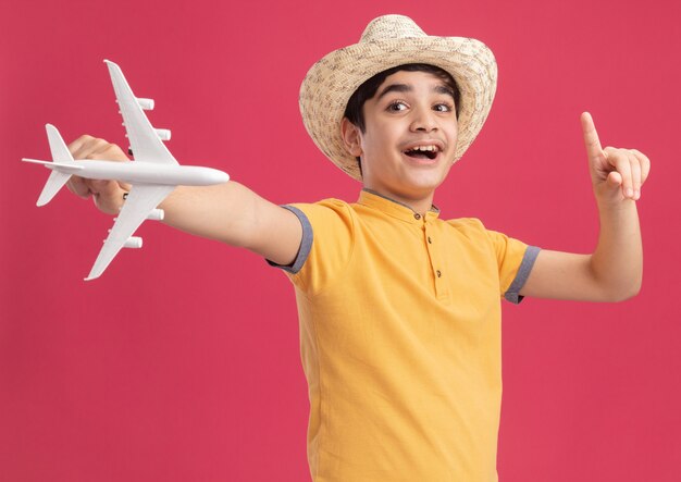 Impressed young caucasian boy wearing beach hat stretching out model plane looking at side pointing up 