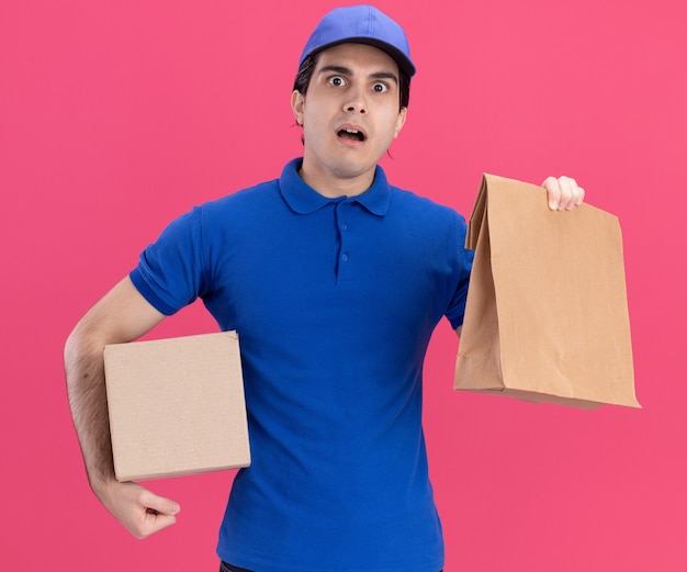 Impressed young caucasian delivery man in blue uniform and cap holding cardbox and paper package 