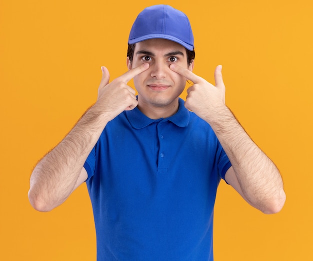 Impressed young caucasian delivery man in blue uniform and cap putting fingers under eyes