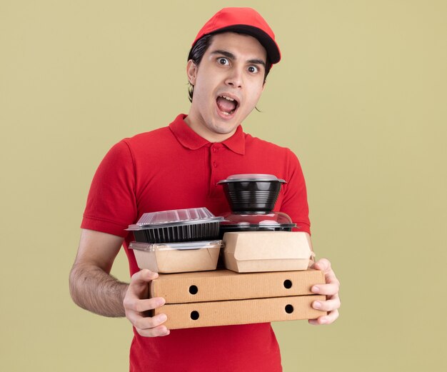 올리브 녹색 벽에 격리된 음식 용기와 종이 음식 패키지가 있는 피자 패키지를 들고 빨간색 유니폼과 모자를 쓴 젊은 백인 배달원