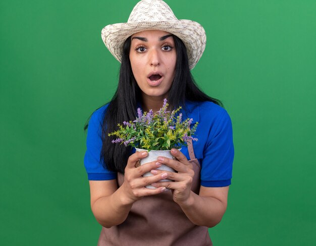 Impressed young caucasian gardener woman wearing uniform and hat holding flowerpot