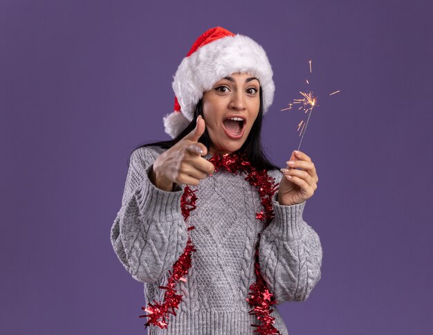 Impressed young caucasian girl wearing christmas hat and tinsel garland around neck holding holiday sparkler isolated on purple wall with copy space
