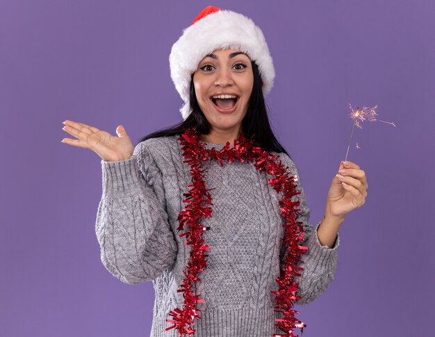 Impressed young caucasian girl wearing christmas hat and tinsel garland around neck holding holiday sparkler looking at camera showing empty hand isolated on purple background