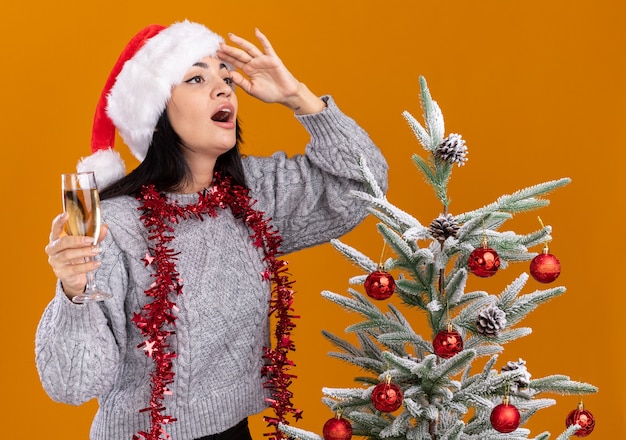 Impressed young caucasian girl wearing christmas hat and tinsel garland around neck standing near decorated christmas tree holding glass of champagne looking at side into distance isolated on orange wall