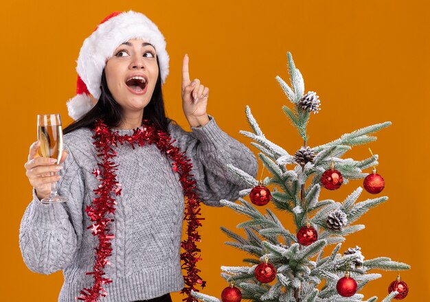impressed young caucasian girl wearing christmas hat and tinsel garland around neck standing near decorated christmas tree holding glass of champagne looking at side pointing up isolated on orange wall