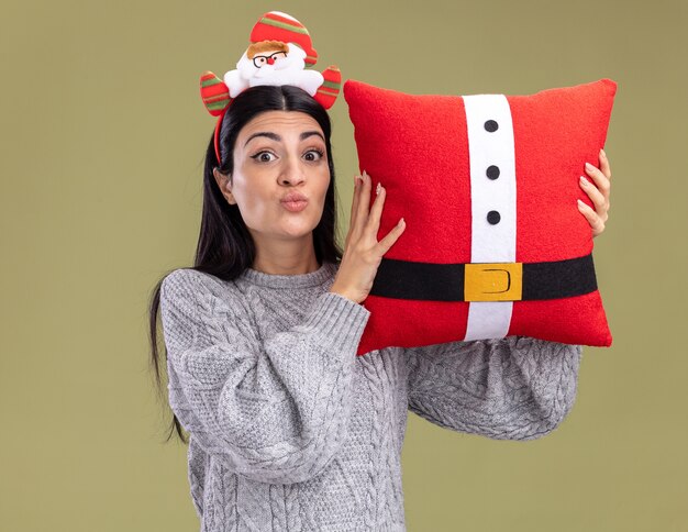 Impressed young caucasian girl wearing santa claus headband holding santa claus pillow near head pursing lips looking at camera isolated on olive green background