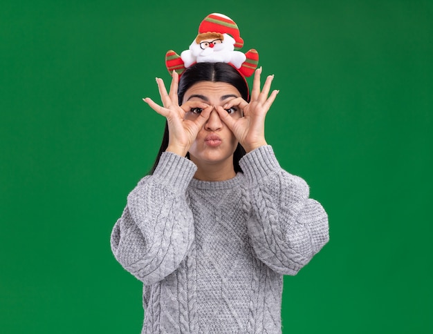 Impressionato giovane ragazza caucasica che indossa la fascia di babbo natale che guarda l'obbiettivo che fa il gesto di sguardo usando le mani come binocolo isolato su priorità bassa verde