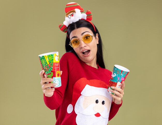 impressed young caucasian girl wearing santa claus headband and sweater with glasses holding plastic christmas cups stretching out one towards  isolated on olive green wall