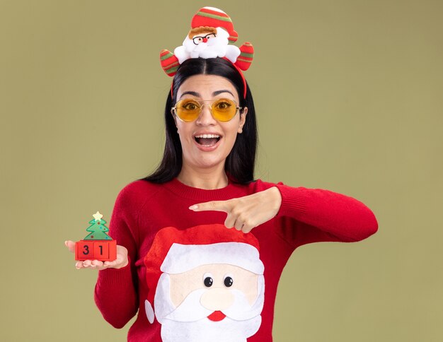 올리브 녹색 벽에 고립 된 날짜와 크리스마스 트리 장난감을 들고 안경을 가리키는 산타 클로스 머리띠와 스웨터를 입고 감동 어린 백인 소녀