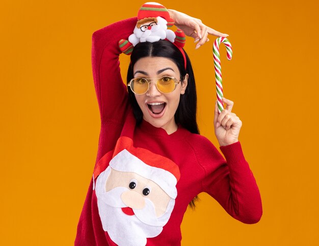 Impressed young caucasian girl wearing santa claus headband and sweater with glasses holding traditional christmas candy cane near head looking at camera isolated on orange background