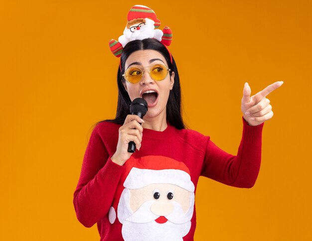 Впечатленная молодая кавказская девушка в головной повязке санта-клауса и свитере в очках разговаривает в микрофон, смотрит и указывает на сторону, изолированную на оранжевой стене
