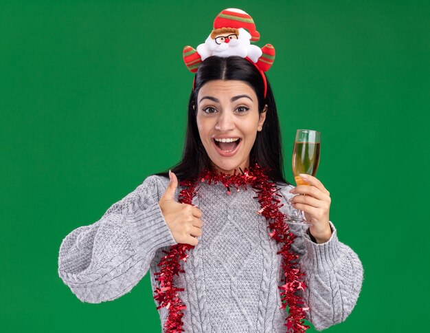 Impressed young caucasian girl wearing santa claus headband and tinsel garland around neck holding glass of champagne looking at camera showing thumb up isolated on green background