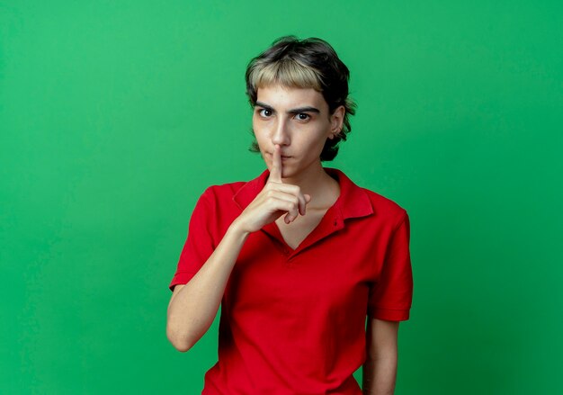 Impressed young caucasian girl with pixie haircut gesturing silence at camera isolated on green background with copy space