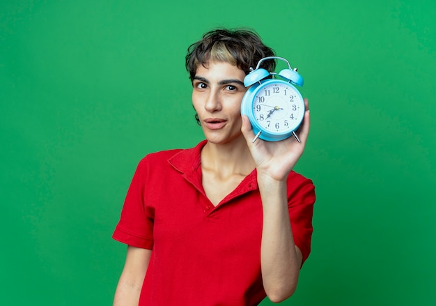 Impressed young caucasian girl with pixie haircut holding alarm clock isolated on green background with copy space