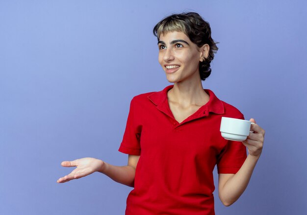 Impressed young caucasian girl with pixie haircut holding cup looking straight and showing empty hand isolated on purple background