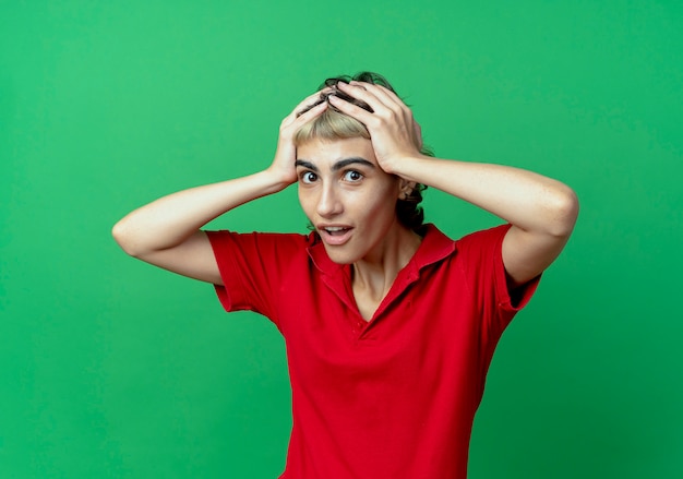 Foto gratuita impressionato giovane ragazza caucasica con taglio di capelli pixie tenendo la sua testa isolata su sfondo verde