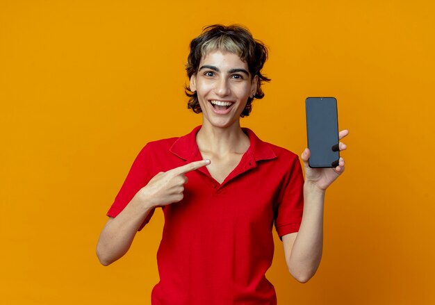 Впечатленная молодая кавказская девушка со стрижкой пикси, держащая и указывающая на мобильный телефон, изолированный на оранжевом фоне с копией пространства