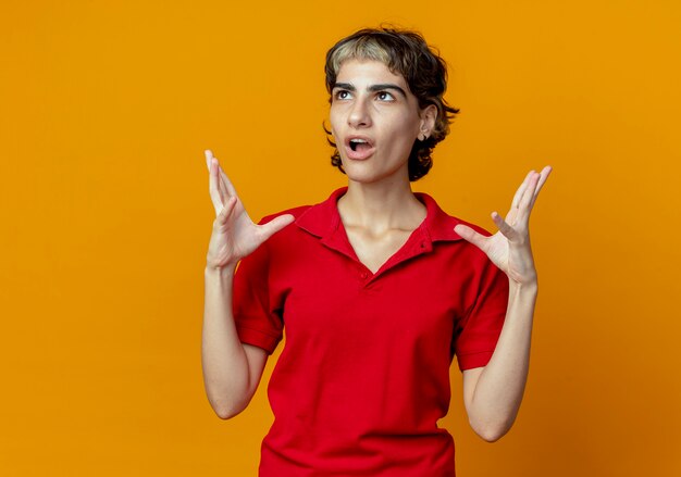 Impressed young caucasian girl with pixie haircut keeping hands in air looking up isolated on orange background with copy space