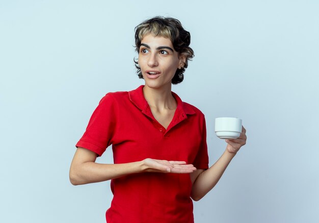 Впечатленная молодая кавказская девушка со стрижкой пикси смотрит в сторону и показывает рукой на чашку, изолированную на белом фоне