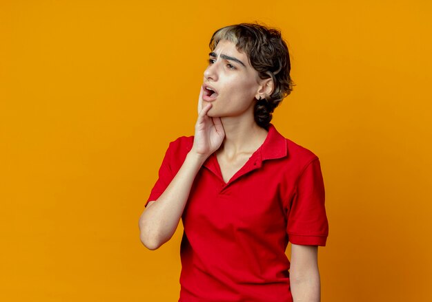 Impressed young caucasian girl with pixie haircut putting hand on chin looking at side isolated on orange background with copy space