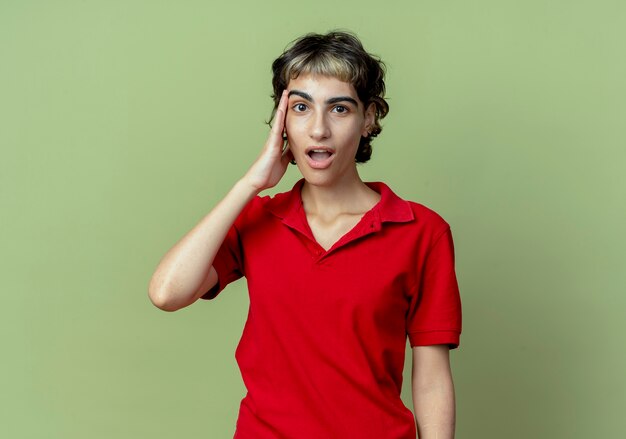 Impressed young caucasian girl with pixie haircut putting hand on temple isolated on olive green background with copy space