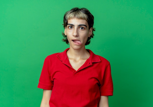 Impressed young caucasian girl with pixie haircut showing tongue looking at camera isolated on green background with copy space