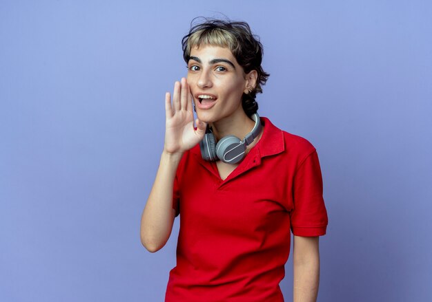 Impressed young caucasian girl with pixie haircut wearing headphones on neck putting hand near mouth whispering at camera isolated on purple background with copy space