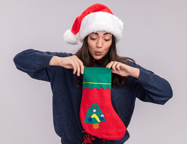 Impressed young caucasian girl with santa hat and garland around neck holds and looks at christmas stocking isolated on white wall with copy space