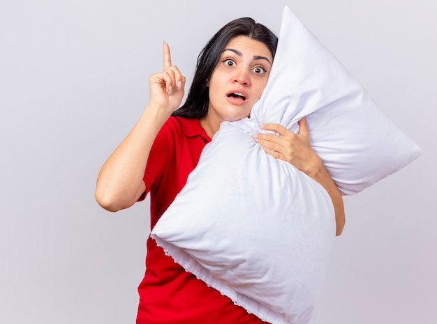 Impressed young caucasian ill girl hugging pillow looking at camera raising finger isolated on white background with copy space