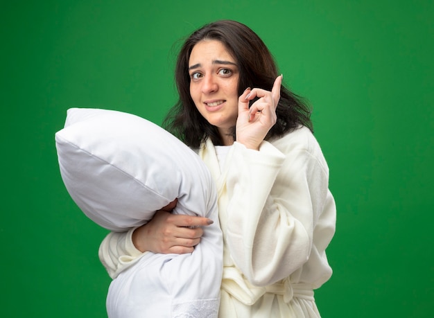 Впечатленная молодая кавказская больная девушка в халате, стоящая в профиль и обнимающая подушку, глядя в камеру, указывая вверх, изолированную на зеленом фоне с копией пространства