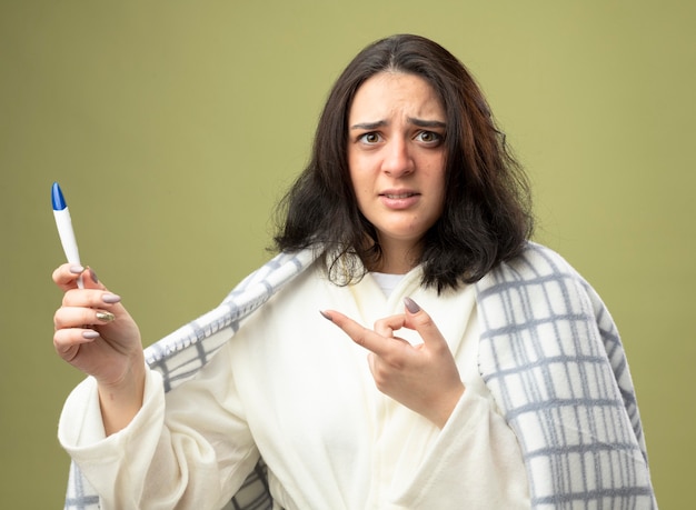 Impressed young caucasian ill girl wearing robe wrapped in plaid holding and pointing at thermometer looking at camera isolated on olive green background