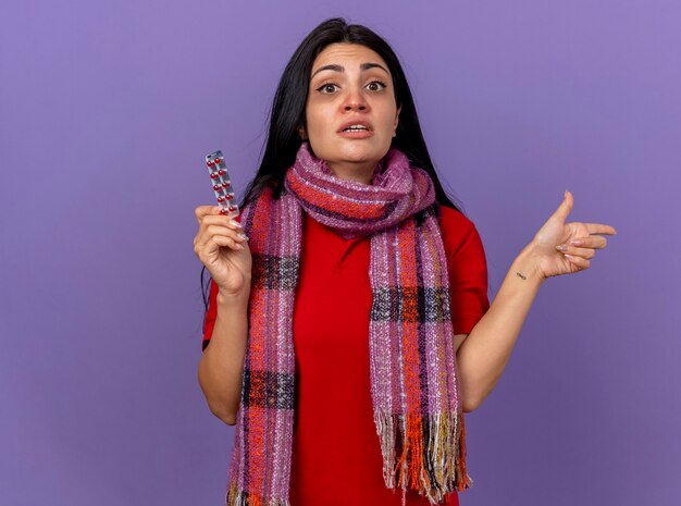 Impressed young caucasian ill girl wearing scarf holding pack of capsules looking at camera pointing at side isolated on purple background with copy space