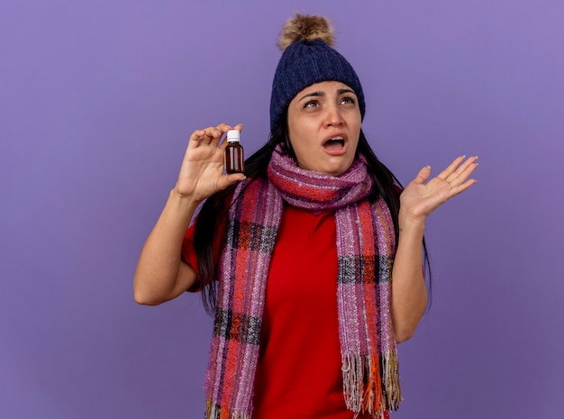 無料写真 冬の帽子とスカーフを身に着けている印象的な若い白人の病気の少女は、コピースペースで紫色の壁に隔離された空の手を見せて見上げるガラスの薬を保持しています
