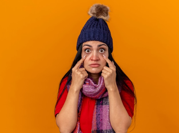 Impressed young caucasian ill girl wearing winter hat and scarf looking at camera pulling down eye lids isolated on orange background with copy space