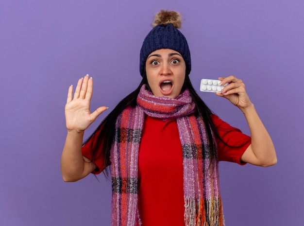 Impressed young caucasian ill girl wearing winter hat and scarf showing pack of tablets looking at camera showing empty hand isolated on purple background