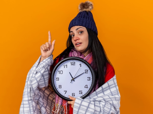 복사 공간 오렌지 배경에 고립 가리키는 카메라를보고 격자 무늬 지주 시계에 싸여 겨울 모자와 스카프를 착용하는 감동 된 젊은 백인 아픈 소녀