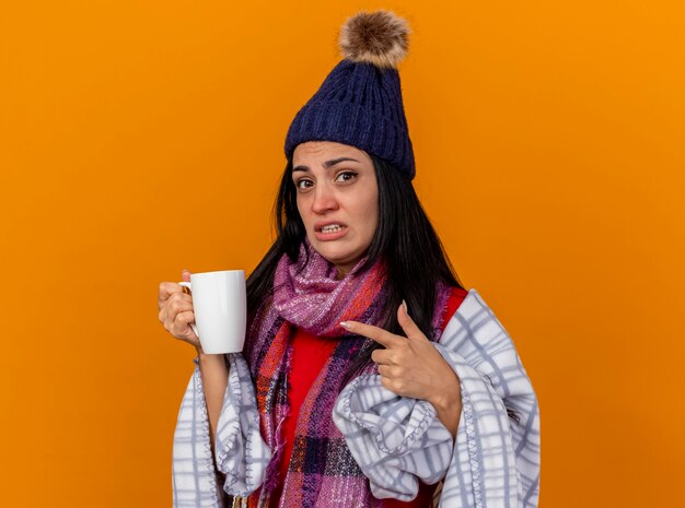 Impressed young caucasian ill girl wearing winter hat and scarf wrapped in plaid holding and pointing at cup of tea looking at camera isolated on orange background with copy space