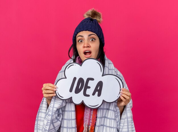 Impressed young caucasian ill girl wearing winter hat and scarf wrapped in plaid looking at camera holding idea bubble isolated on crimson background with copy space