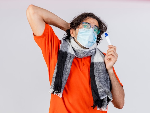 Impressionato giovane indoeuropeo uomo malato con gli occhiali e maschera tenendo il termometro guardando la telecamera tenendo la mano dietro la testa isolata su sfondo bianco