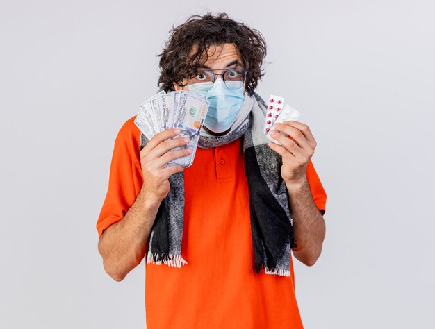 Impressed young caucasian ill man wearing glasses scarf and mask holding money and medical pills looking at camera isolated on white background with copy space