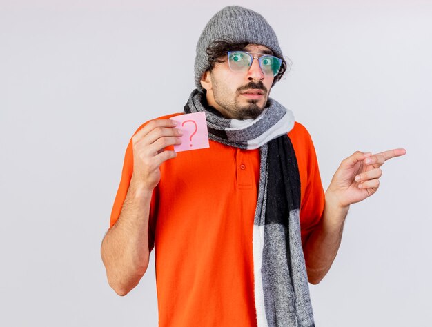 Impressed young caucasian ill man wearing glasses winter hat and scarf holding question note looking at camera pointing at side isolated on white background with copy space