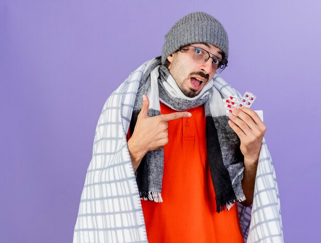 Impressed young caucasian ill man wearing glasses winter hat and scarf wrapped in plaid holding and pointing at packs of capsules  isolated on purple wall with copy space